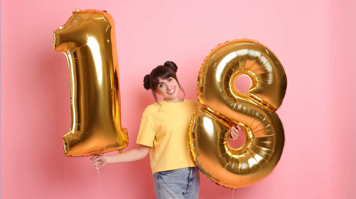 Uśmiechnięta dziewczyna trzymająca złote balony w kształcie liczby 18, ubrana w żółtą koszulkę i dżinsy, na różowym tle
