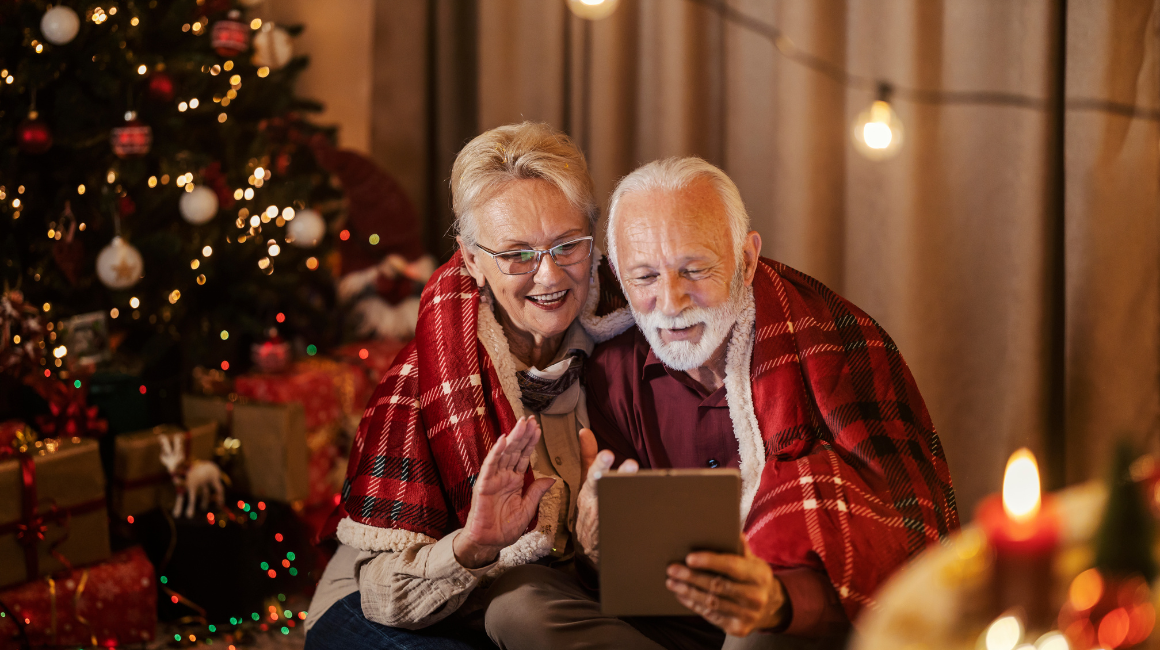 Uśmiechnięci seniorzy pod kocem w kratę przy choince rozmawiają przez tablet w świątecznej atmosferze